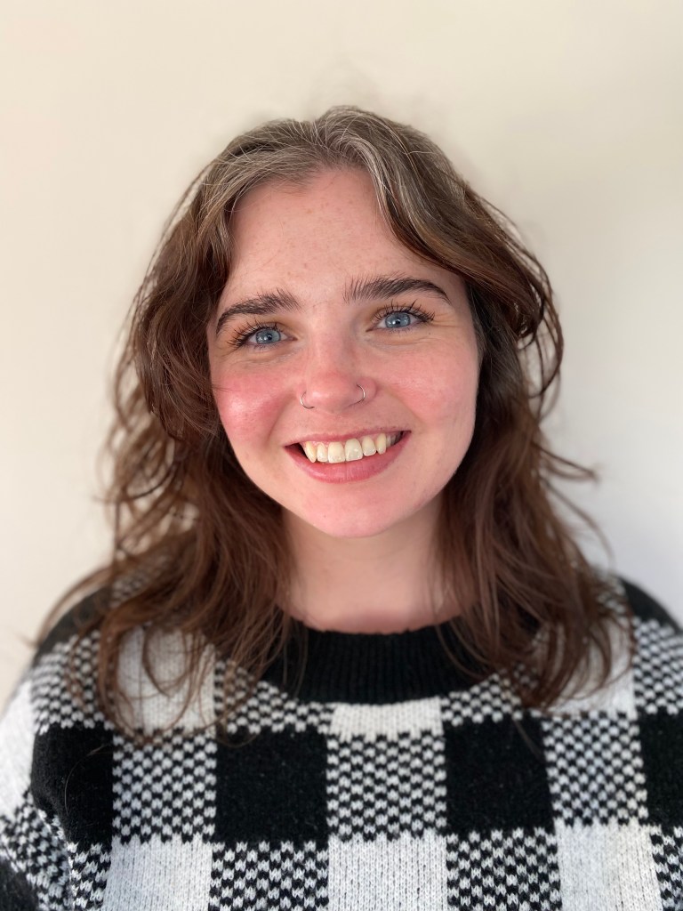 A close-up photo of a white woman with wavy medium-length brown hair. She has blue eyes and two silver nose hoop piercings. She is smiling and wearing a black and white checkered sweater.